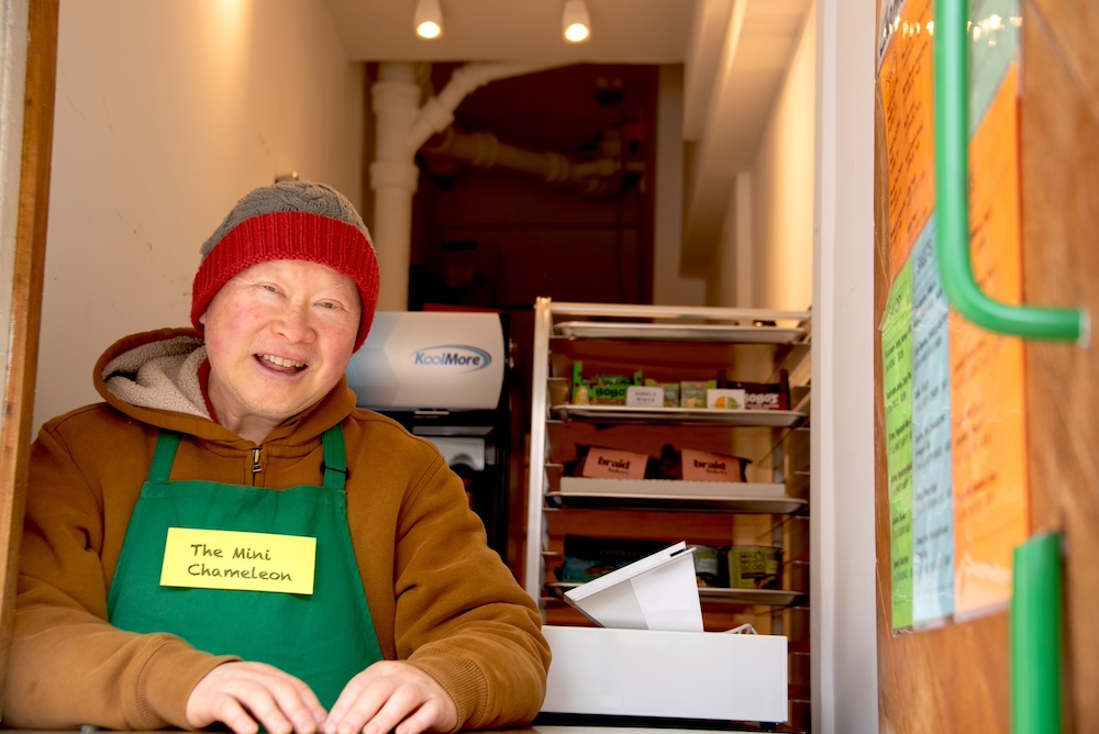 Darryl Dea, a middle-aged Chinese American man wearing an ochre hoodie, red and grey beanie, and green apron, poses inside his snack kiosk and smiles for the camera.