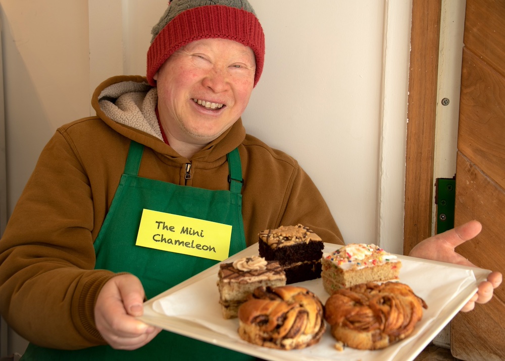 Darryl Dea, a middle-aged Chinese American man wearing an ochre hoodie, red and grey beanie, and green apron, holds a plate of pastries and smiles for the camera.