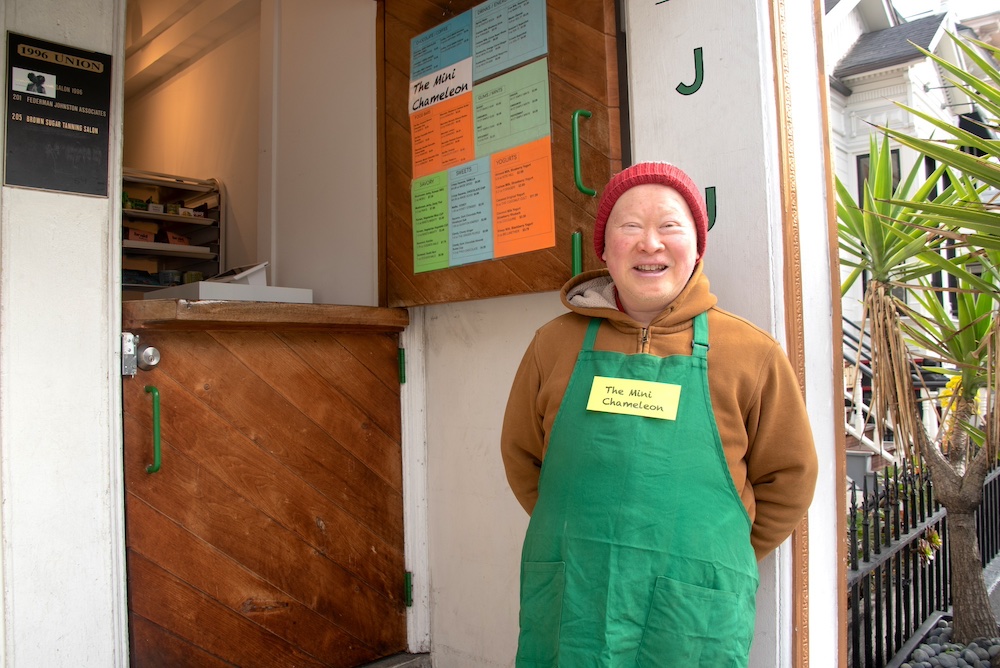 Darryl Dea, a middle-aged Chinese American man wearing an ochre hoodie, red and grey beanie, and green apron, stands outside his snack kiosk menu and smiles for the camera.