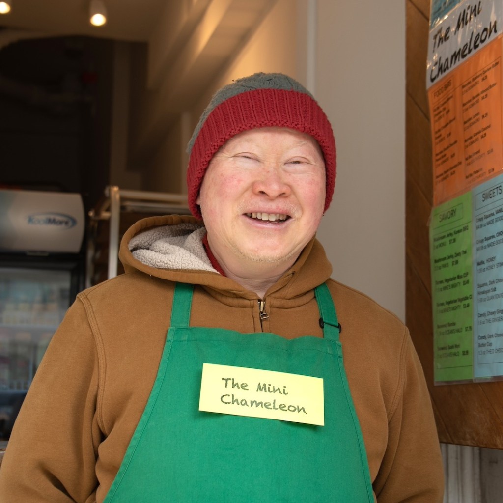 Darryl Dea, a middle-aged Chinese American man wearing an ochre hoodie, red and grey beanie, and green apron, stands next to his snack kiosk menu and smiles for the camera.