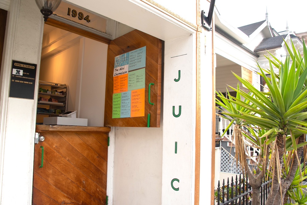 The Mini Chameleon snack shack exterior, a small wooden countertop and menu display framed by white exterior walls with the letters JUIC painted in the doorway and the addreess, 1994, is visible above.